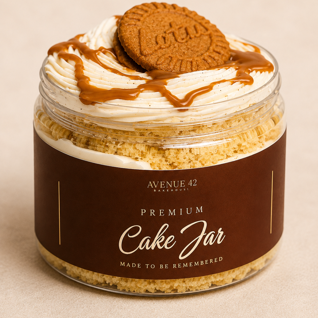 A clear jar filled with a layered cake, topped with whipped cream, caramel drizzle, and a round cookie, with a dark brown label reading 'PREMIUM Cake Jar' and 'AVENUE 42 BAKERY'.