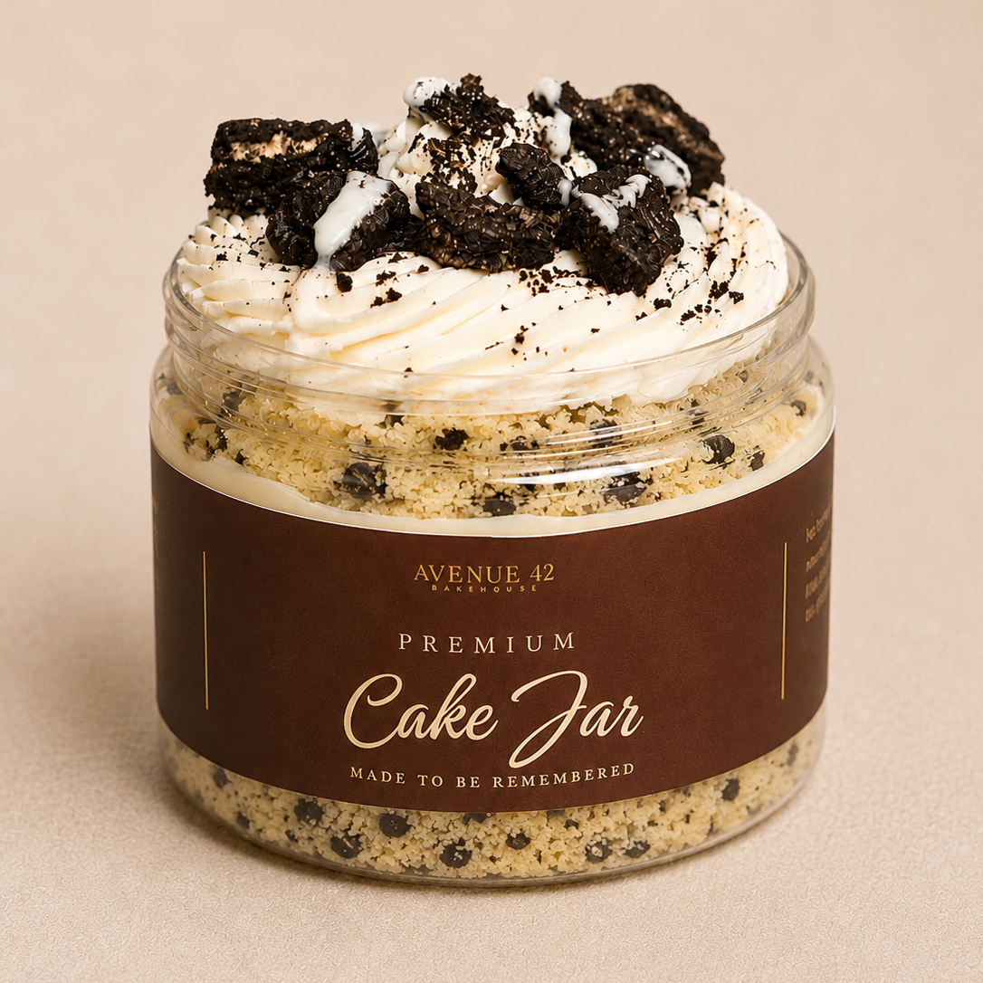 A clear jar filled with a layered dessert cake, topped with whipped cream, cookie crumbs, and dark cookie chunks, with a brown label reading 'PREMIUM Cake Jar' on a beige background.