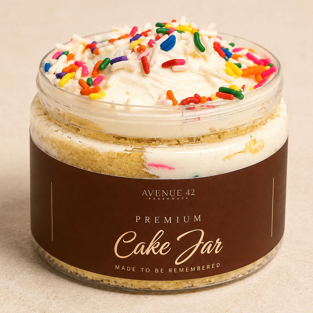 A clear jar filled with a layered cake and topped with whipped cream and colorful sprinkles, wrapped in a brown paper label that reads 'PREMIUM Cake Jar' by Avenue 42 Bakehouse, on a neutral beige background.