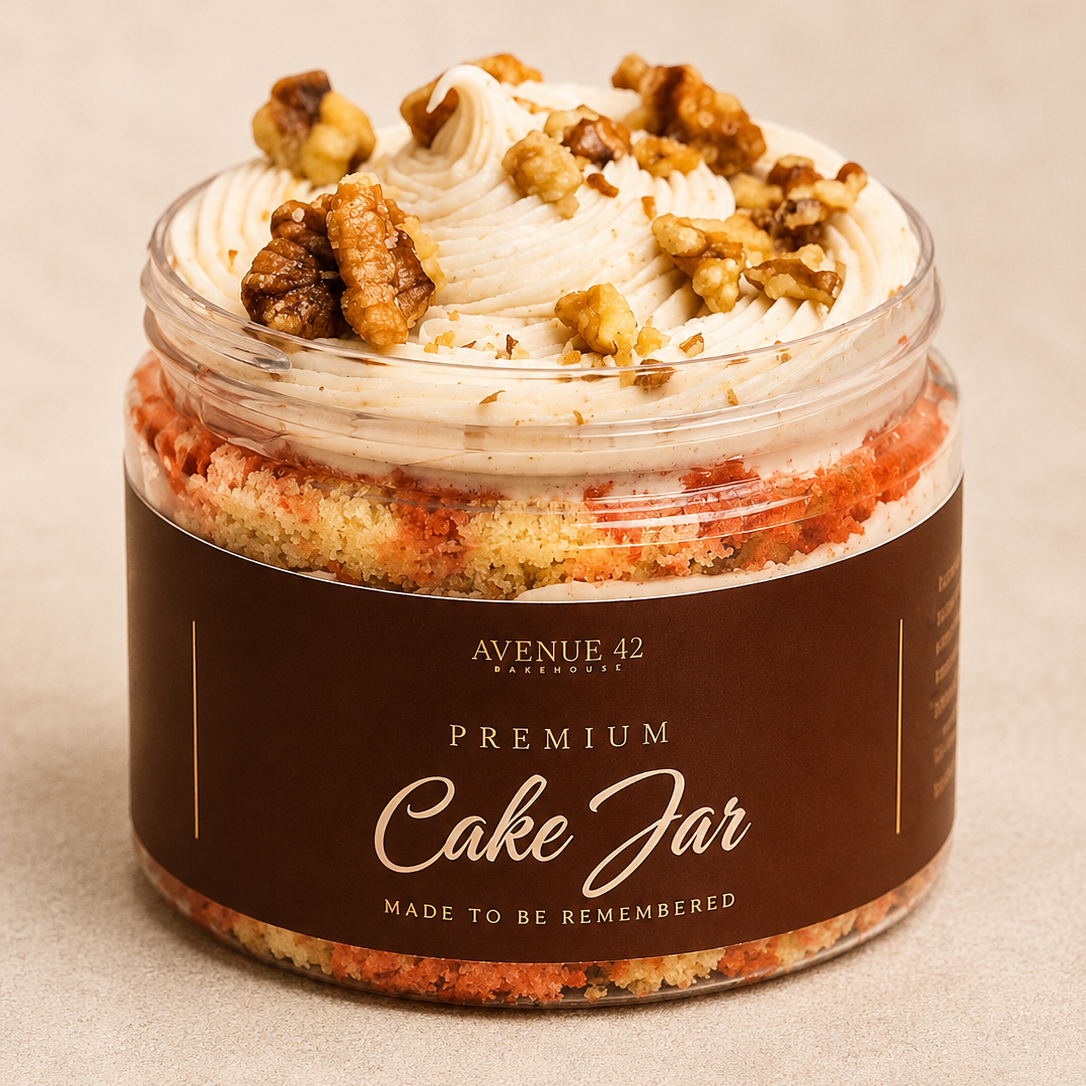 A jar filled with a layered carrot cake, topped with buttercream and chopped walnuts, wrapped in a dark brown label that reads 'PREMIUM Cake Jar' by Avenue 42 Bakery.