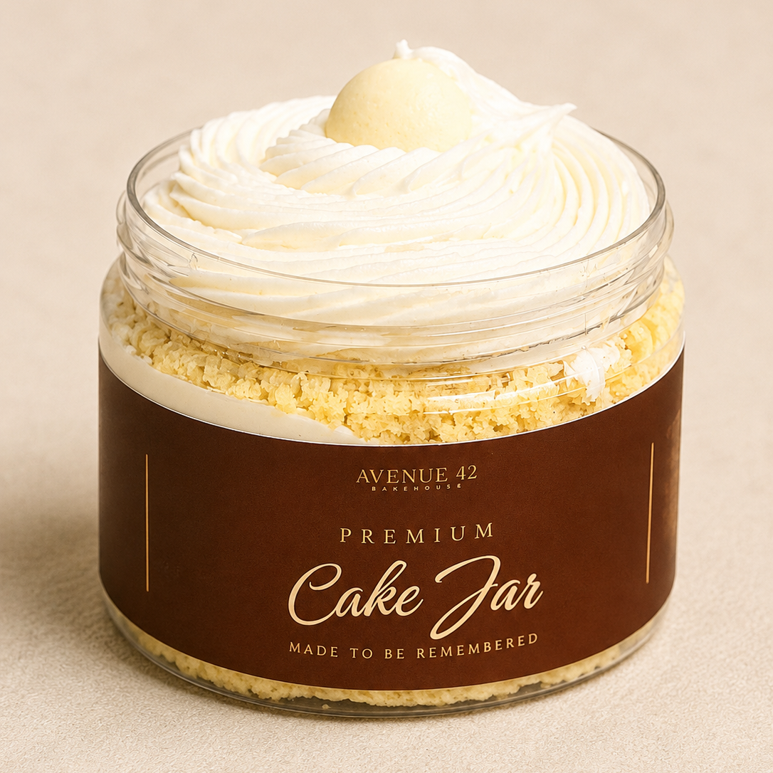 A clear jar filled with a cream-colored cake, topped with whipped cream and a small white chocolate ball. The jar has a dark brown paper label reading 'PREMIUM Cake Jar' and 'AVENUE 42 Bakehouse'.